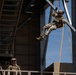 Two military personnel are engaged in a rappelling or rope descent training exercise at what appears to be a dedicated training facility. One soldier is suspended mid-air on orange/yellow ropes attached to a steel tower structure, while a second soldier observes from a platform below. Both are wearing full combat gear including helmets, body armor, and camouflage uniforms.