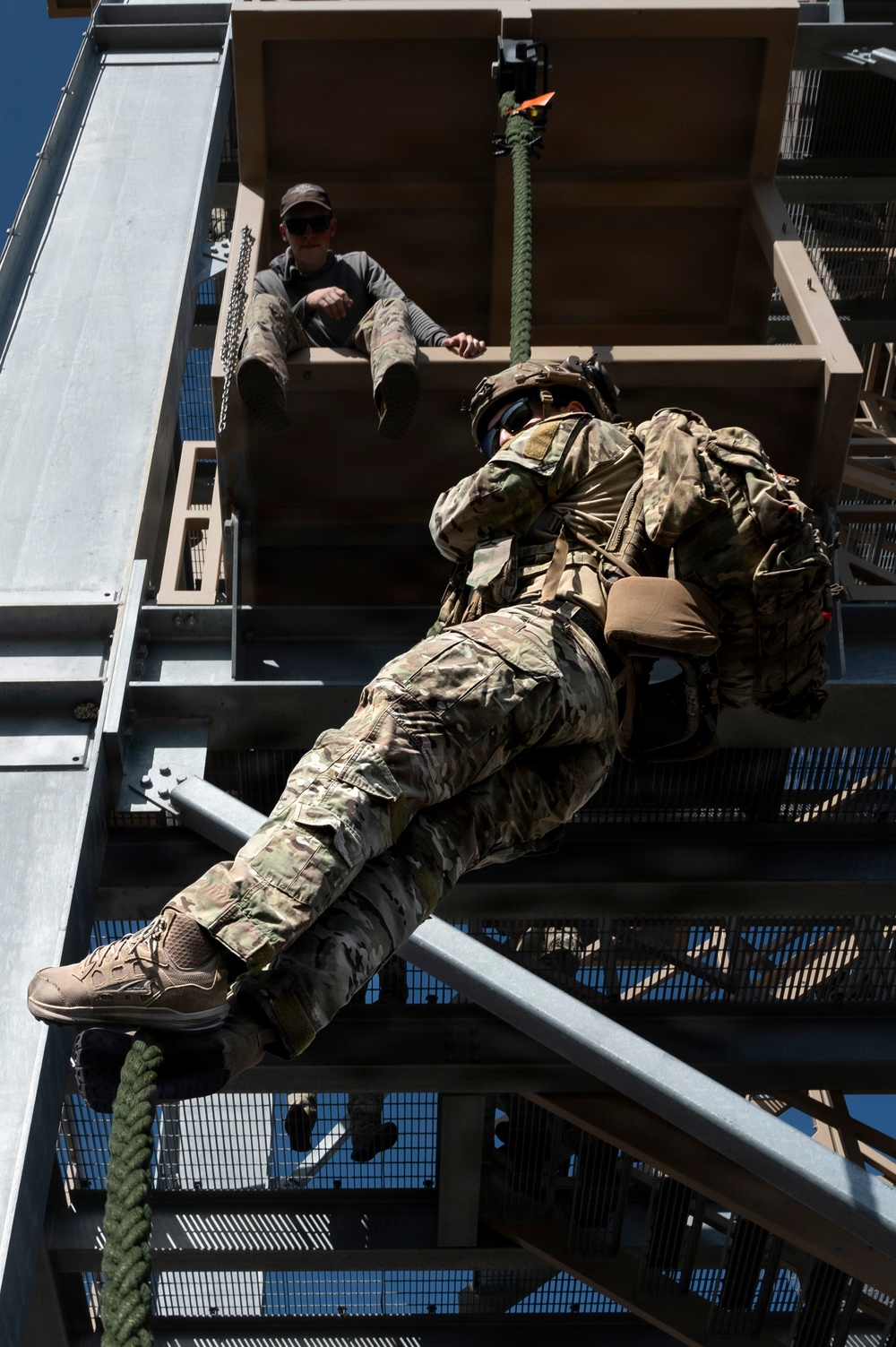 A military or special operations personnel in full combat gear, including helmet, body armor, and large rucksack, is rappelling down a training tower using a rope. An instructor or observer in civilian-style clothing sits at the top of the tower monitoring the descent. The image is taken from a low angle looking upward, emphasizing the height and scale of the training structure.