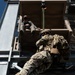 A military or special operations personnel in full combat gear, including helmet, body armor, and large rucksack, is rappelling down a training tower using a rope. An instructor or observer in civilian-style clothing sits at the top of the tower monitoring the descent. The image is taken from a low angle looking upward, emphasizing the height and scale of the training structure.