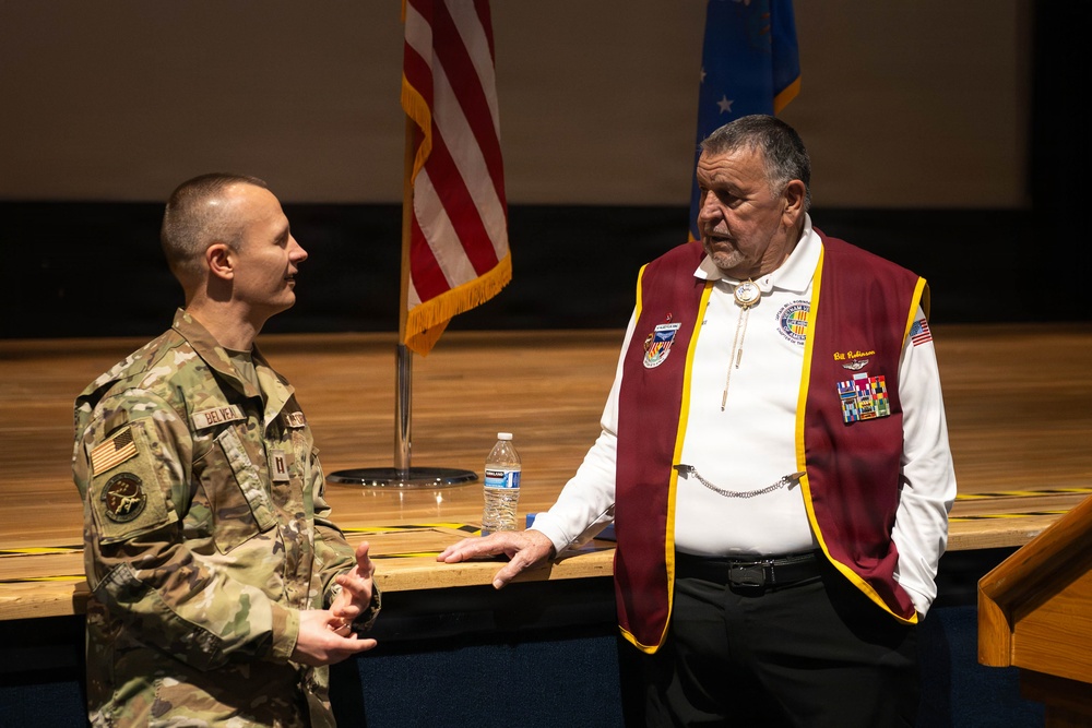 A U.S. Army soldier in multicam/OCP uniform is engaged in conversation with an older civilian man wearing a maroon and gold veteran's organization jacket adorned with multiple pins and medals. The setting appears to be an auditorium or ceremonial hall, with U.S. and Air Force flags visible in the background. The interaction appears informal and friendly, likely occurring before or after an official event.
