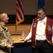A U.S. Army soldier in multicam/OCP uniform is engaged in conversation with an older civilian man wearing a maroon and gold veteran's organization jacket adorned with multiple pins and medals. The setting appears to be an auditorium or ceremonial hall, with U.S. and Air Force flags visible in the background. The interaction appears informal and friendly, likely occurring before or after an official event.