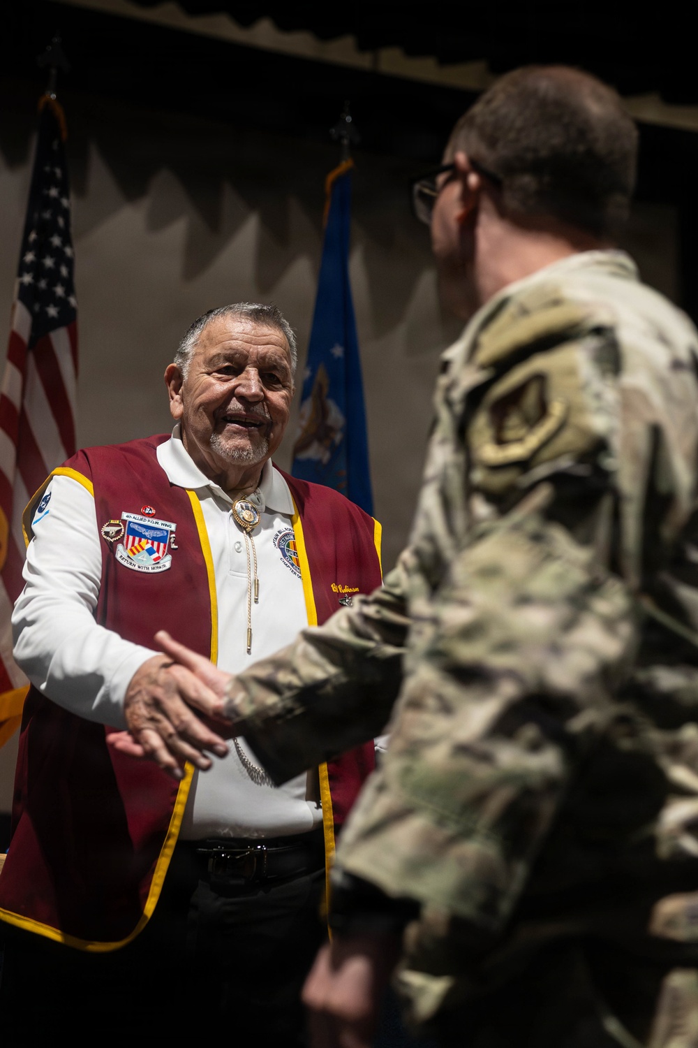 An older civilian man wearing a maroon and gold vest with military organization patches is shaking hands with a uniformed military service member in camouflage (OCP/MultiCam) pattern. The setting appears to be a formal military ceremony or recognition event, with American and Air Force flags visible in the background. The civilian's vest displays patches including what appears to be '4th Allied POW Wing' and 'Return with Honor' insignia.