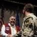 An older civilian man wearing a maroon and gold vest with military organization patches is shaking hands with a uniformed military service member in camouflage (OCP/MultiCam) pattern. The setting appears to be a formal military ceremony or recognition event, with American and Air Force flags visible in the background. The civilian's vest displays patches including what appears to be '4th Allied POW Wing' and 'Return with Honor' insignia.