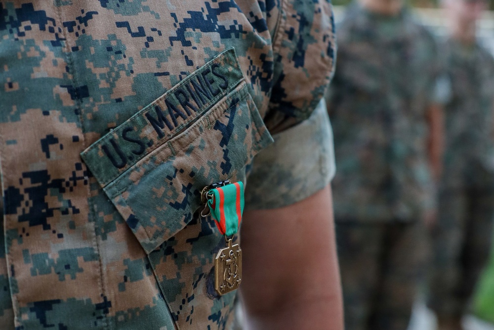 A close-up photograph of a U.S. Marines service member wearing the Marine Pattern (MARPAT) digital camouflage uniform. A military medal with a green and red striped ribbon is pinned to the chest pocket of the uniform. Additional uniformed personnel are visible but out of focus in the background.