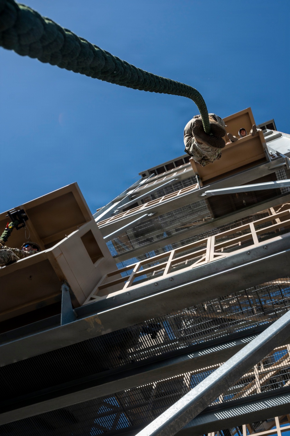 A dramatic low-angle upward shot of a military rappelling tower, with a thick braided rope extending from the foreground toward the top of the structure. At least one person in military/camouflage uniform is visible at the top platform preparing to rappel, while another uniformed individual is visible on a lower platform. The tower features multiple levels with metal grating stairs and wooden platform sections against a clear blue sky.