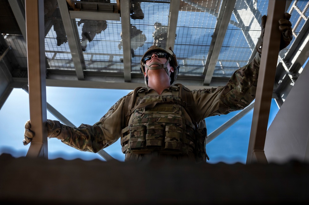 A military service member in full tactical gear is photographed from a low-angle perspective, looking upward while gripping metal railings or bars on either side. The dramatic upward shot captures the soldier against a blue sky backdrop with a metal grated structure overhead. The composition emphasizes the soldier's tactical equipment and purposeful posture.