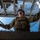 A military service member in full tactical gear is photographed from a low-angle perspective, looking upward while gripping metal railings or bars on either side. The dramatic upward shot captures the soldier against a blue sky backdrop with a metal grated structure overhead. The composition emphasizes the soldier's tactical equipment and purposeful posture.