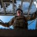 A military service member in full tactical gear is photographed from a low-angle perspective, looking upward while gripping metal railings or bars on either side. The dramatic upward shot captures the soldier against a blue sky backdrop with a metal grated structure overhead. The composition emphasizes the soldier's tactical equipment and purposeful posture.