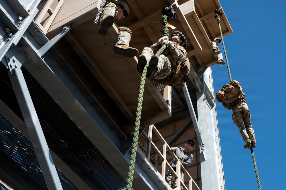 Military personnel in camouflage uniforms are conducting rappelling or fast-rope training exercises from an elevated training tower structure. Multiple soldiers are visible at various stages of descending ropes, while an instructor in civilian clothing observes from a lower platform. The image is taken from a low angle looking upward, emphasizing the height and dynamic nature of the training activity.