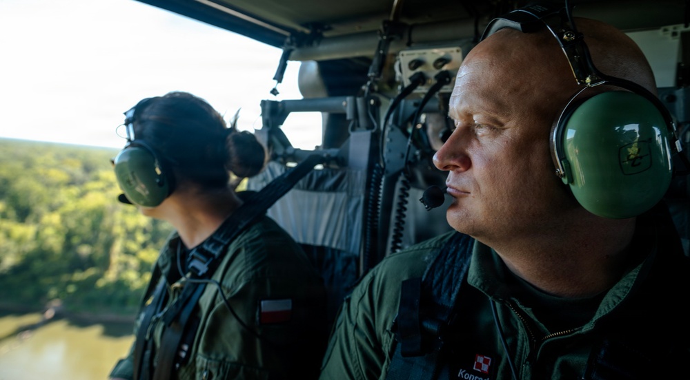 Two military personnel wearing green flight suits and aviation headsets are seated inside a military helicopter in flight. Both individuals display Polish national flag patches on their uniforms, indicating they are members of the Polish Armed Forces. The helicopter is flying over a forested area with a visible river or waterway below.