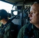 Two military personnel wearing green flight suits and aviation headsets are seated inside a military helicopter in flight. Both individuals display Polish national flag patches on their uniforms, indicating they are members of the Polish Armed Forces. The helicopter is flying over a forested area with a visible river or waterway below.