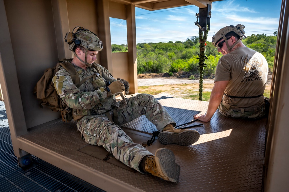 Two military personnel are seated on a diamond-plate metal platform inside what appears to be a rappelling or fast-rope training tower. One individual is in full combat gear including multicam uniform, tactical vest, backpack, and helmet with communications equipment, while the other wears a tan t-shirt, camouflage pants, and a helmet with sunglasses. A rope or cable is visible hanging from the structure, suggesting this is a height/rappel training facility.
