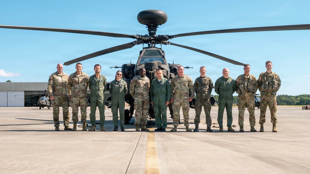 A group of approximately eleven military personnel pose for a formal group photograph on an airfield tarmac in front of an AH-64 Apache attack helicopter. Personnel are wearing a mix of camouflage (OCP/MultiCam) uniforms and olive drab flight suits, suggesting a mix of ground and aviation crew members. The setting appears to be a military airbase with hangars and additional helicopters visible in the background.