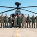 A group of approximately eleven military personnel pose for a formal group photograph on an airfield tarmac in front of an AH-64 Apache attack helicopter. Personnel are wearing a mix of camouflage (OCP/MultiCam) uniforms and olive drab flight suits, suggesting a mix of ground and aviation crew members. The setting appears to be a military airbase with hangars and additional helicopters visible in the background.