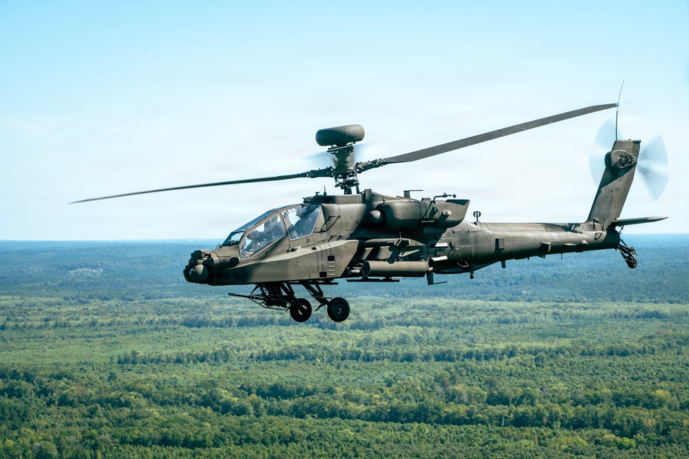 A military attack helicopter is captured in flight over a dense forested landscape under a clear sky. The aircraft is photographed from a slightly elevated angle, showing its full profile in sharp detail. The helicopter appears to be on a routine flight or training mission.