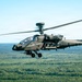 A military attack helicopter is captured in flight over a dense forested landscape under a clear sky. The aircraft is photographed from a slightly elevated angle, showing its full profile in sharp detail. The helicopter appears to be on a routine flight or training mission.