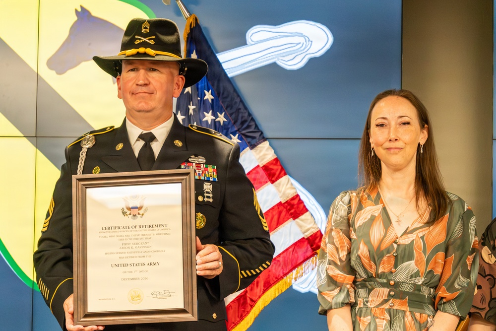 A U.S. Army First Sergeant in dress uniform holds a Certificate of Retirement from the United States Army, dated December 2026, standing beside a woman in a floral dress. The ceremony appears to be a military retirement event, with an American flag and what appears to be a 1st Cavalry Division backdrop visible behind them. The soldier is wearing a cavalry Stetson hat with crossed sabers insignia and multiple service ribbons.