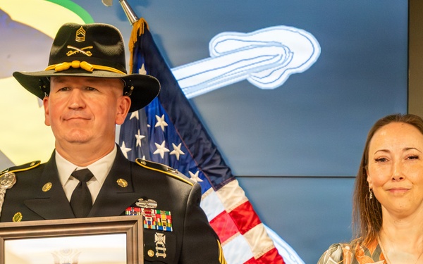 A U.S. Army First Sergeant in dress uniform holds a Certificate of Retirement from the United States Army, dated December 2026, standing beside a woman in a floral dress. The ceremony appears to be a military retirement event, with an American flag and what appears to be a 1st Cavalry Division backdrop visible behind them. The soldier is wearing a cavalry Stetson hat with crossed sabers insignia and multiple service ribbons.
