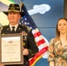A U.S. Army First Sergeant in dress uniform holds a Certificate of Retirement from the United States Army, dated December 2026, standing beside a woman in a floral dress. The ceremony appears to be a military retirement event, with an American flag and what appears to be a 1st Cavalry Division backdrop visible behind them. The soldier is wearing a cavalry Stetson hat with crossed sabers insignia and multiple service ribbons.