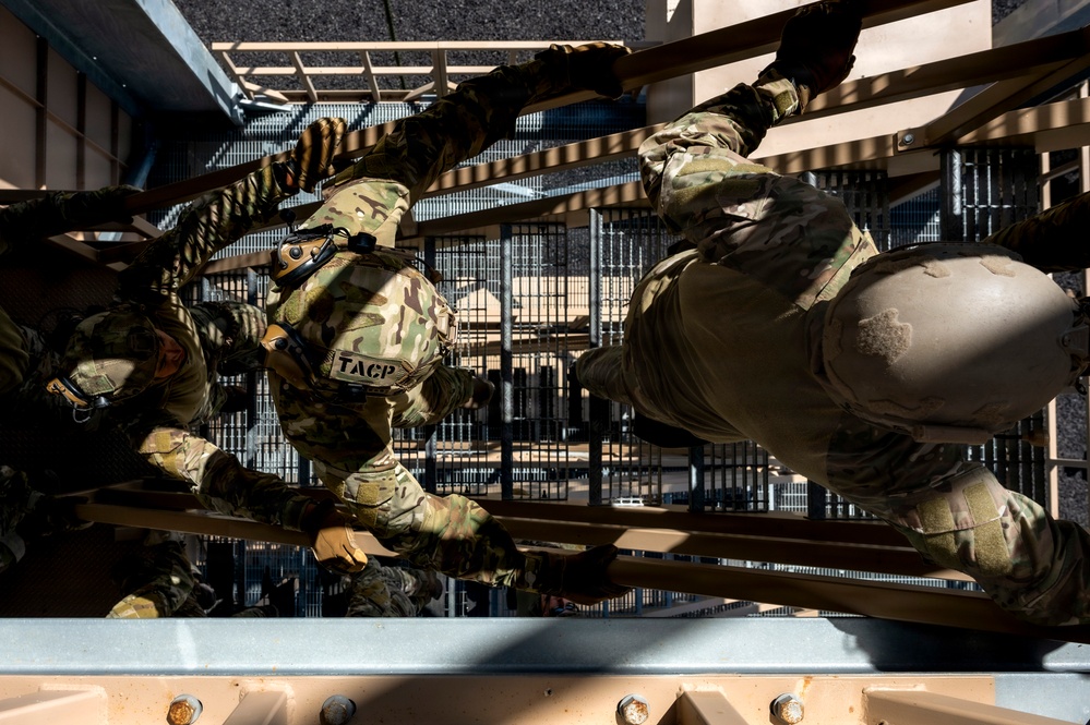 Military personnel in MultiCam/OCP camouflage uniforms are conducting a physical training or obstacle course exercise, climbing over or traversing a structure from an overhead perspective. At least one individual has a visible 'TACP' (Tactical Air Control Party) patch, identifying them as U.S. Air Force special operations personnel. The image is taken from directly above, creating a dynamic top-down composition.