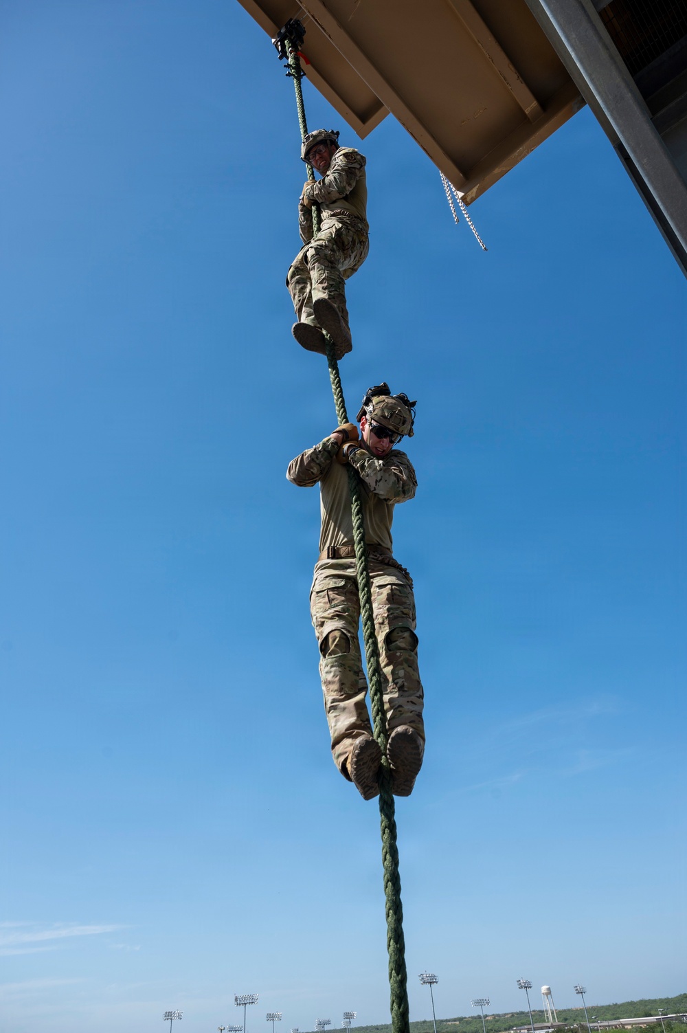 Two military personnel in camouflage uniforms are climbing a green rope suspended from an elevated structure, photographed from a low angle looking upward. The image captures a rope climbing exercise or training drill, with one soldier higher on the rope and another below. A stadium with light towers and a water tower are visible in the distant background.