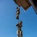 Two military personnel in camouflage uniforms are climbing a green rope suspended from an elevated structure, photographed from a low angle looking upward. The image captures a rope climbing exercise or training drill, with one soldier higher on the rope and another below. A stadium with light towers and a water tower are visible in the distant background.