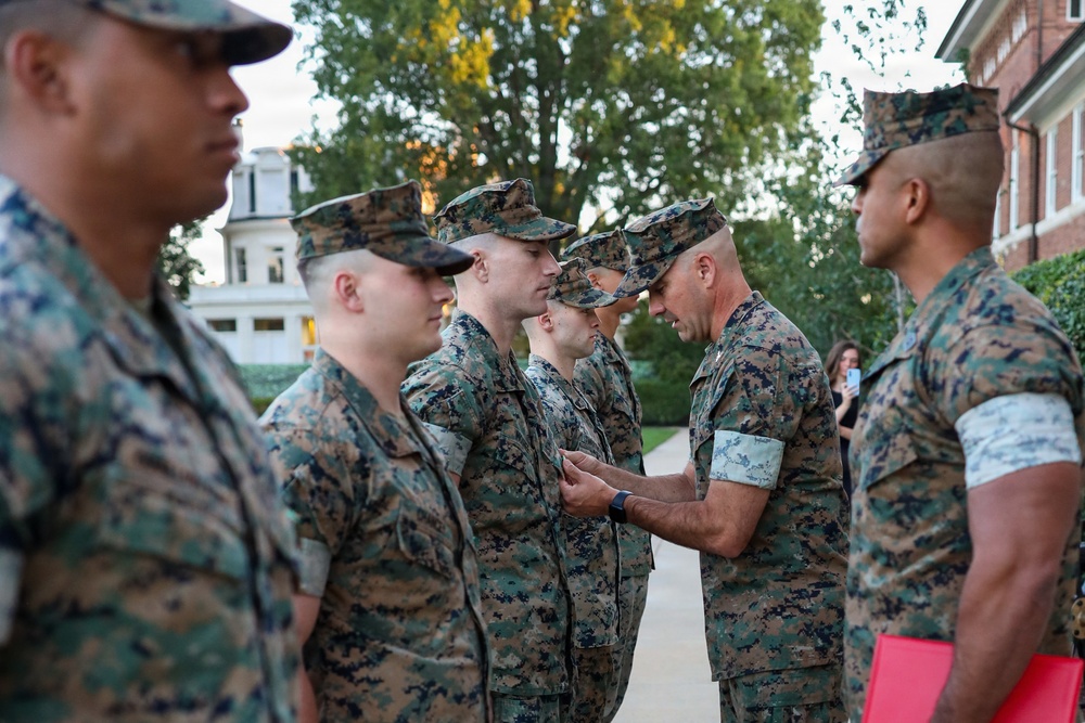 A military ceremony is taking place outdoors, likely a decoration or award presentation. A senior Marine in MARPAT camouflage uniform is pinning a medal or device onto the chest of another Marine while others stand in formation. The setting appears to be a formal military installation with brick buildings and trees visible in the background.