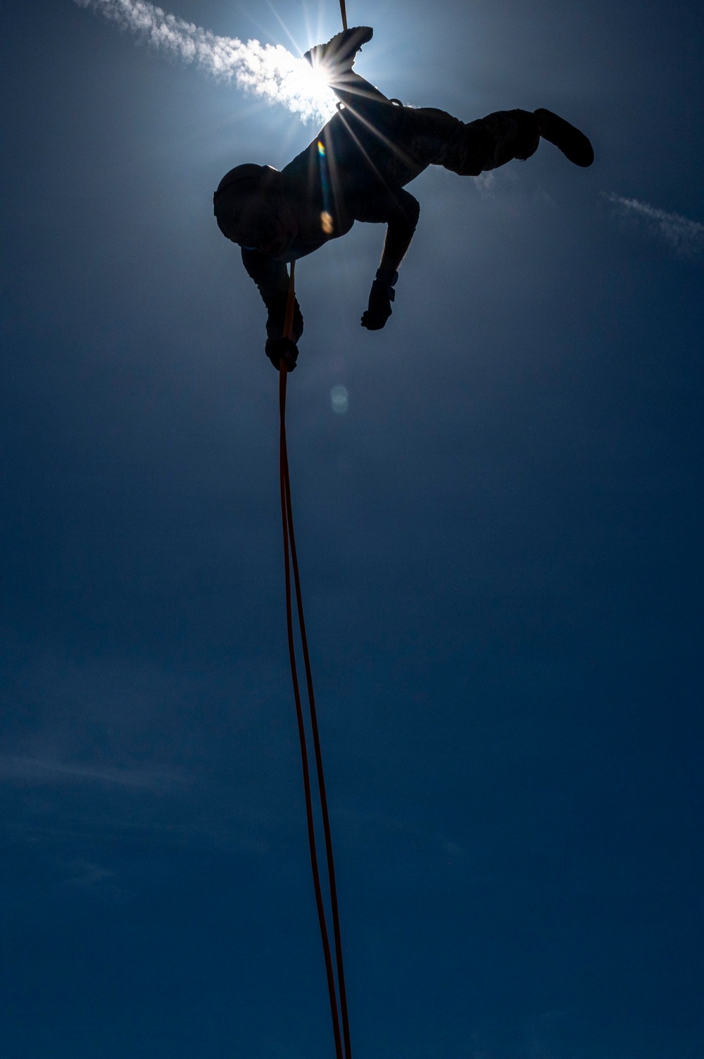 A silhouetted person is suspended upside-down or at an inverted angle from a rope against a bright blue sky, with the sun creating a strong starburst lens flare directly behind them. The figure appears to be rappelling or performing a rope descent technique, holding what appears to be an orange/red rope. The dramatic low-angle, backlit composition creates a striking contrast between the dark human silhouette and the luminous sky.