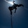 A silhouetted person is suspended upside-down or at an inverted angle from a rope against a bright blue sky, with the sun creating a strong starburst lens flare directly behind them. The figure appears to be rappelling or performing a rope descent technique, holding what appears to be an orange/red rope. The dramatic low-angle, backlit composition creates a striking contrast between the dark human silhouette and the luminous sky.