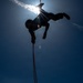 A silhouetted person is suspended upside-down or at an inverted angle from a rope against a bright blue sky, with the sun creating a strong starburst lens flare directly behind them. The figure appears to be rappelling or performing a rope descent technique, holding what appears to be an orange/red rope. The dramatic low-angle, backlit composition creates a striking contrast between the dark human silhouette and the luminous sky.