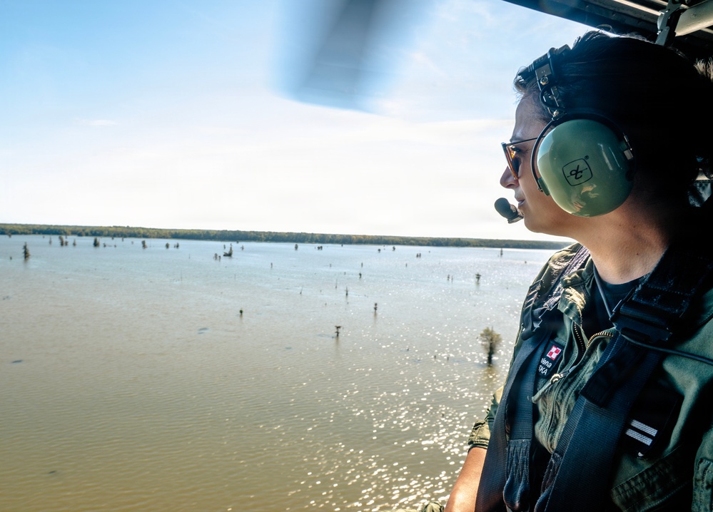 A person wearing military or emergency response gear, green aviation headset, sunglasses, and a tactical vest with a red cross patch is observing a flooded landscape from an open helicopter door. The aerial view reveals a wide expanse of brown floodwater with partially submerged trees visible across the area. This appears to be a flood assessment or disaster response aerial survey mission.