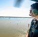 A person wearing military or emergency response gear, green aviation headset, sunglasses, and a tactical vest with a red cross patch is observing a flooded landscape from an open helicopter door. The aerial view reveals a wide expanse of brown floodwater with partially submerged trees visible across the area. This appears to be a flood assessment or disaster response aerial survey mission.