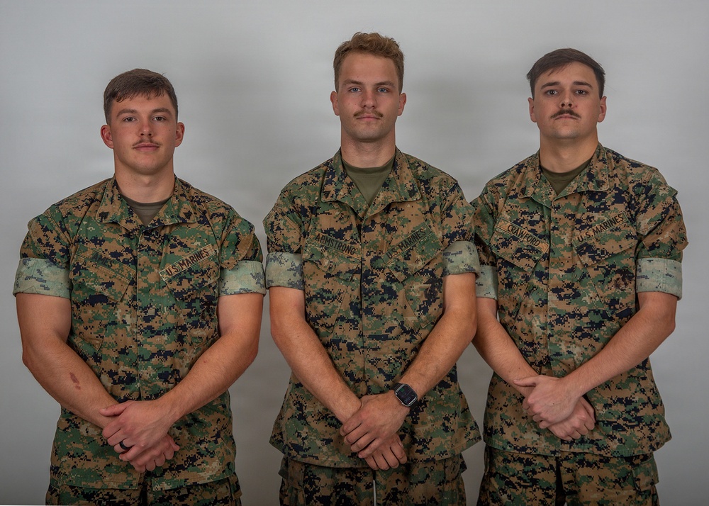 Three U.S. Marines in MARPAT woodland camouflage uniforms pose for a formal group portrait against a neutral gray background. Their name tapes are visible, identifying them as Armstrong (center) and Crawford (right); the left individual's name is partially obscured. All three stand in a relaxed parade-rest-style stance with hands clasped in front.