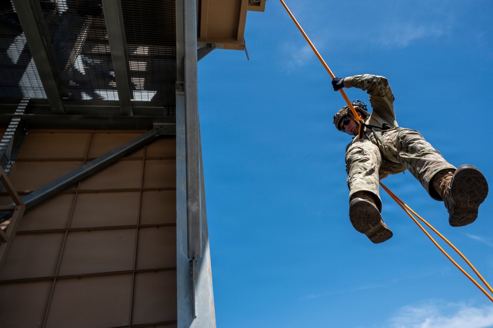 A military or law enforcement personnel is rappelling down the side of a training tower using orange ropes and a harness system. The individual is wearing full camouflage uniform, tactical boots, gloves, a helmet, and eye protection. The photo is taken from a low angle looking upward, emphasizing the height and dynamic nature of the descent.