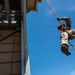 A military or law enforcement personnel is rappelling down the side of a training tower using orange ropes and a harness system. The individual is wearing full camouflage uniform, tactical boots, gloves, a helmet, and eye protection. The photo is taken from a low angle looking upward, emphasizing the height and dynamic nature of the descent.