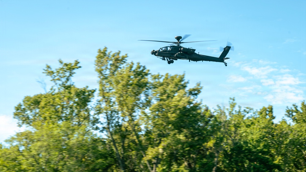A military attack helicopter, identifiable as an AH-64 Apache, is flying low over a treeline against a partly cloudy blue sky. The aircraft appears to be in active flight, flying at low altitude just above the canopy of green deciduous trees. The image was taken from a ground-level perspective looking upward.
