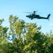 A military attack helicopter, identifiable as an AH-64 Apache, is flying low over a treeline against a partly cloudy blue sky. The aircraft appears to be in active flight, flying at low altitude just above the canopy of green deciduous trees. The image was taken from a ground-level perspective looking upward.