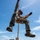 A military service member in full camouflage uniform is rappelling or performing a rope descent exercise using a bright orange rope. The individual is suspended mid-air against a blue sky, with a wooded landscape and a multi-story building visible in the background. The subject appears to be smiling, suggesting a training exercise rather than a combat scenario.