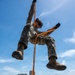 A military service member in full camouflage uniform is rappelling or performing a rope descent exercise using a bright orange rope. The individual is suspended mid-air against a blue sky, with a wooded landscape and a multi-story building visible in the background. The subject appears to be smiling, suggesting a training exercise rather than a combat scenario.