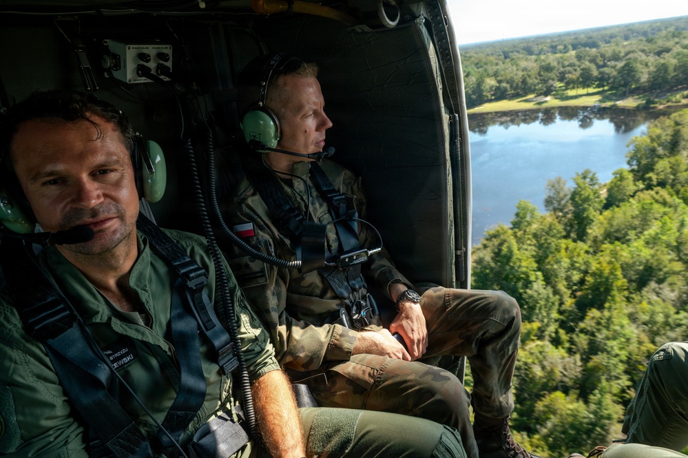 Two military personnel are seated inside an open-door helicopter in flight, observing the landscape below. The individual on the right wears a Polish flag patch on their uniform, suggesting Polish military affiliation. The aerial view reveals a forested area with a river or lake visible below.