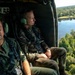 Two military personnel are seated inside an open-door helicopter in flight, observing the landscape below. The individual on the right wears a Polish flag patch on their uniform, suggesting Polish military affiliation. The aerial view reveals a forested area with a river or lake visible below.