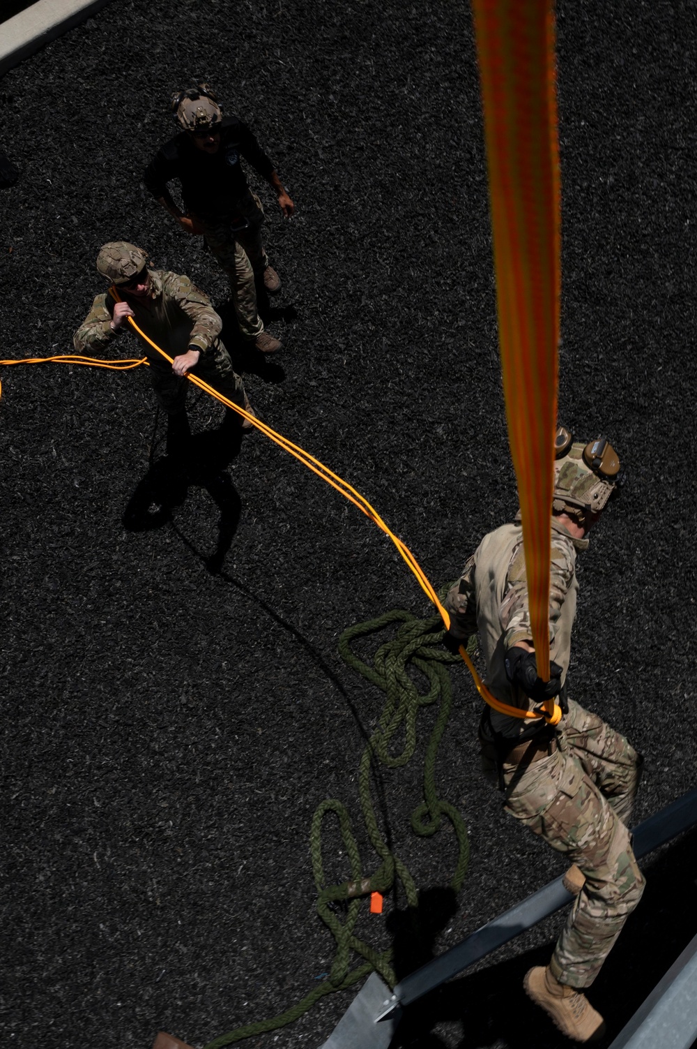 An aerial top-down view of military personnel conducting a rappelling or rope descent training exercise. One soldier is positioned at the edge of a structure holding an orange rappelling rope/strap, while two others on the ground manage orange guide ropes and a coiled green rope. The image is taken from an elevated vantage point, likely from the top of the training structure.
