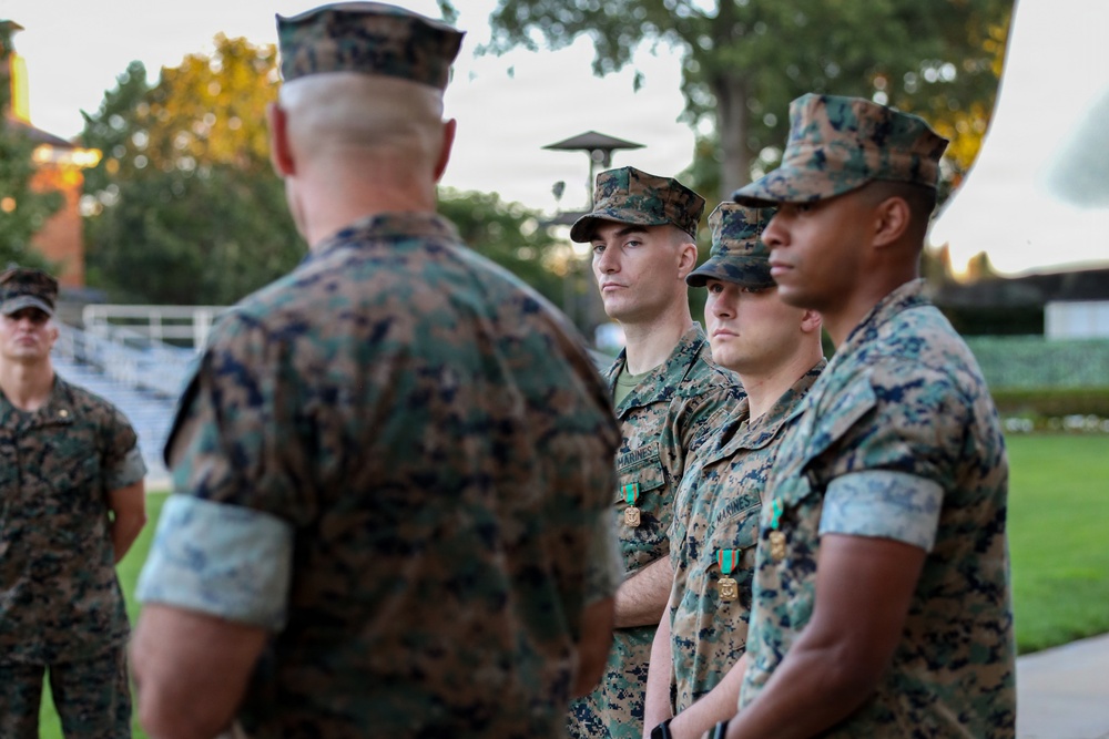 A military awards or recognition ceremony involving U.S. Marines in woodland MARPAT camouflage uniforms. Three Marines standing in a line are being addressed by a senior Marine whose back is to the camera. At least two of the standing Marines are visibly wearing medals with green and gold ribbons pinned to their uniforms.