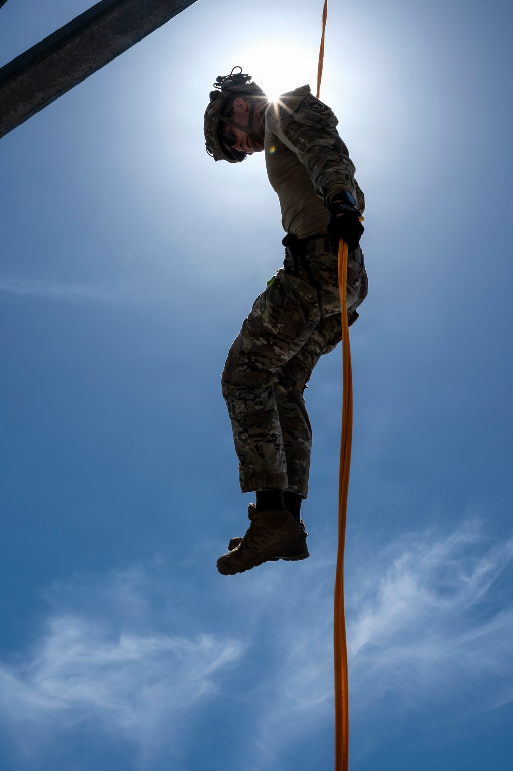 A military personnel in full camouflage uniform is rappelling or descending on an orange rope, photographed from a low angle looking upward against a bright blue sky. The sun creates a starburst effect directly behind the subject, producing a dramatic silhouette effect. The image captures a training or operational descent exercise from an elevated structure.