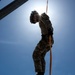 A military personnel in full camouflage uniform is rappelling or descending on an orange rope, photographed from a low angle looking upward against a bright blue sky. The sun creates a starburst effect directly behind the subject, producing a dramatic silhouette effect. The image captures a training or operational descent exercise from an elevated structure.