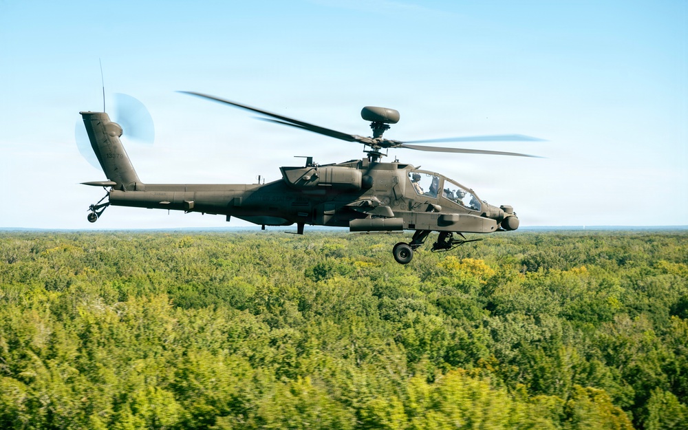 A military attack helicopter, identifiable as a Boeing AH-64 Apache, is captured in flight at low altitude over a dense green forest canopy. The helicopter is flying in a level attitude with its rotor blades in motion, visible as a blur. The image is taken from another airborne platform, suggesting an air-to-air photography session.