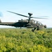 A military attack helicopter, identifiable as a Boeing AH-64 Apache, is captured in flight at low altitude over a dense green forest canopy. The helicopter is flying in a level attitude with its rotor blades in motion, visible as a blur. The image is taken from another airborne platform, suggesting an air-to-air photography session.
