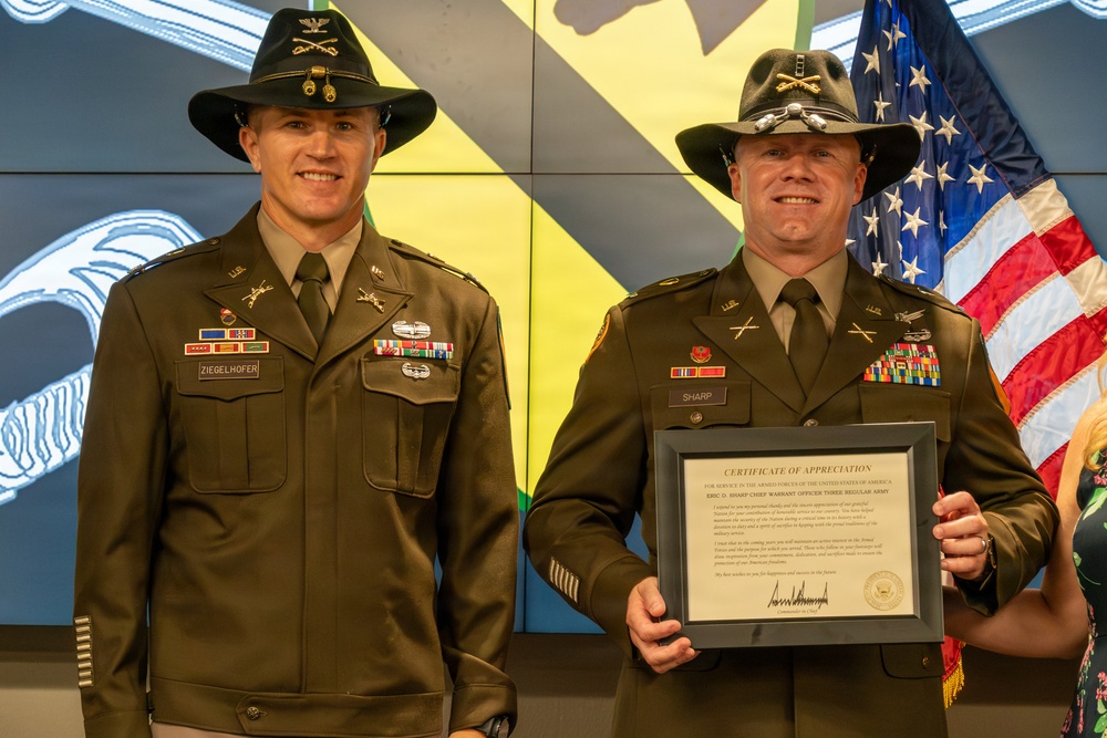 Two U.S. Army soldiers in dress green uniforms and cavalry Stetson hats pose together at what appears to be a military ceremony. The soldier on the right, identified by his name tape as 'SHARP,' is holding a framed Certificate of Appreciation. An American flag is visible in the background along with a digital display showing military insignia.