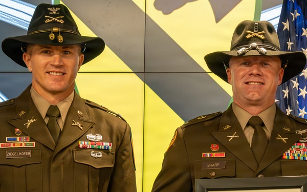 Two U.S. Army soldiers in dress green uniforms and cavalry Stetson hats pose together at what appears to be a military ceremony. The soldier on the right, identified by his name tape as 'SHARP,' is holding a framed Certificate of Appreciation. An American flag is visible in the background along with a digital display showing military insignia.
