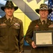 Two U.S. Army soldiers in dress green uniforms and cavalry Stetson hats pose together at what appears to be a military ceremony. The soldier on the right, identified by his name tape as 'SHARP,' is holding a framed Certificate of Appreciation. An American flag is visible in the background along with a digital display showing military insignia.